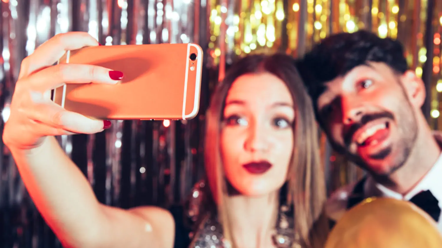Couple en tenue de soirée prenant un selfie devant un rideau à paillettes dorées